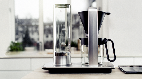 A modern coffee brewing station with a sleek design is displayed on a kitchen counter. The setup includes a clear cylindrical water reservoir to the left, a metal and black coffee filter holder in the center, and a glass carafe with a handle to the right. The background features a blurred view of a window, suggesting this is in a home setting. The focus on the coffee equipment highlights its contemporary aesthetic and suggests an interest in specialty coffee preparation.